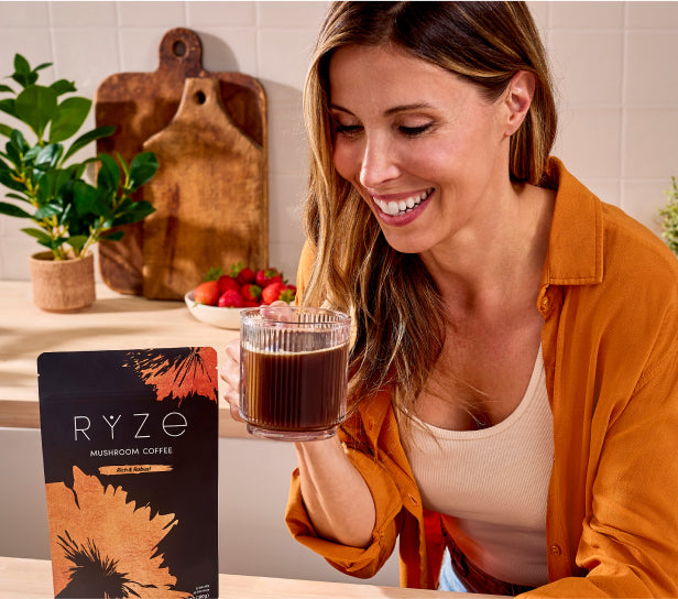 Woman in orange shirt holding a glass of RYZE Mushroom Coffee in a bright kitchen