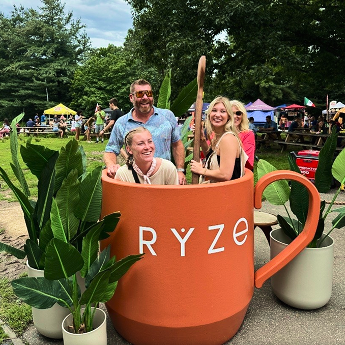 People posing inside a large orange mug with 'RYZE' branding in an outdoor setting.